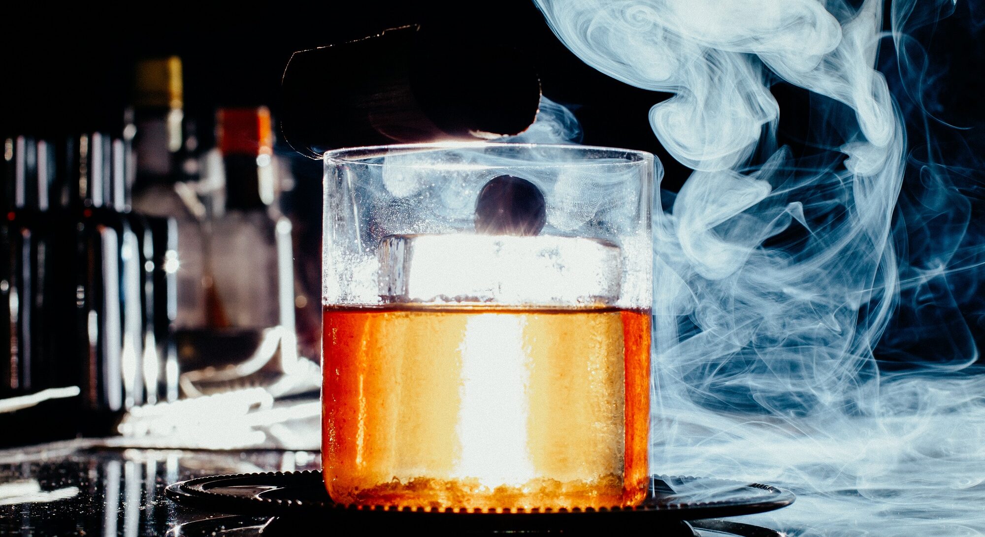 Imagem de um copo com whisky e gelo, com fumaça por cima, ao lado de outros recipientes, em um ambiente com iluminação baixa.