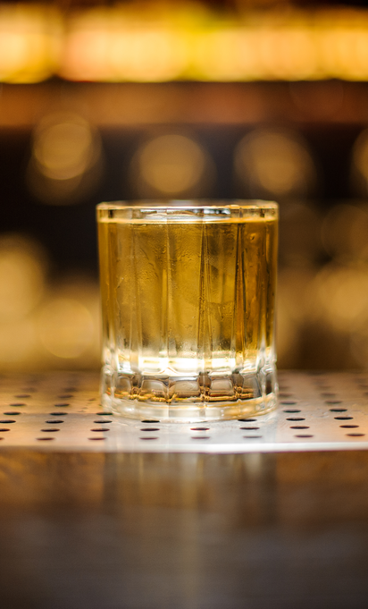Imagem de um copo de vidro com o drink Rusty Nail sobre a bancada de um bar, com objetos desfocados ao fundo.